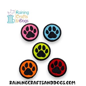 Puede incluir: Cinco parches redondos con huellas de patas bordadas. Cada parche tiene una huella de pata negra sobre un fondo de color: rosa, azul, amarillo, naranja y rojo. También se ve el texto "Raining Crafts & Dogs" y "Rainingcraftsanddogs.com".