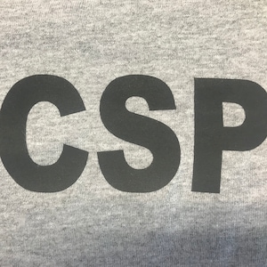 May include: Close-up of a gray t-shirt with the letters "CSP" printed in black. The letters are large and centered on the shirt. The fabric has a textured appearance.