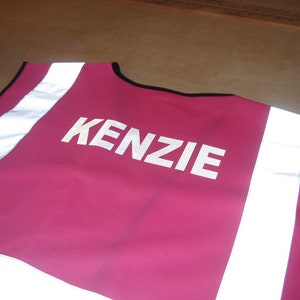 May include: A magenta safety vest with the name "KENZIE" printed in white block letters. The vest has reflective white stripes down the shoulders and black trim around the neckline.