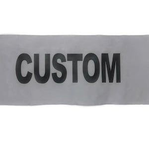 May include: A rectangular, light gray fabric patch with the word "CUSTOM" printed in bold, black letters across the center. The patch has a slightly wrinkled texture and is set against a white background.