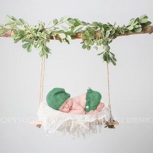 May include: A newborn baby wearing a green knitted hat and matching pants is sleeping on a white fabric swing hanging from a branch with green leaves.