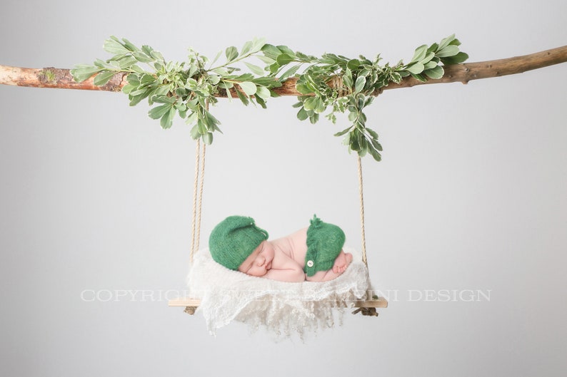 May include: A newborn baby wearing a green knitted hat and matching pants is sleeping on a white fabric swing hanging from a branch with green leaves.