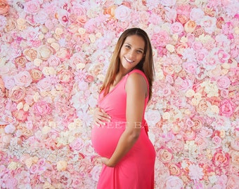 May include: A pregnant woman wearing a pink dress stands in front of a wall of pink and white flowers.