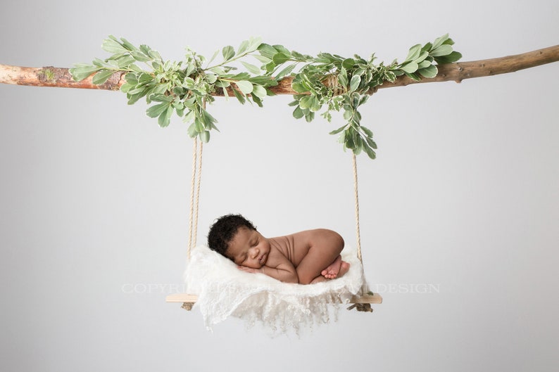 May include: A newborn baby sleeping on a white blanket in a wooden swing hanging from a branch with green leaves.