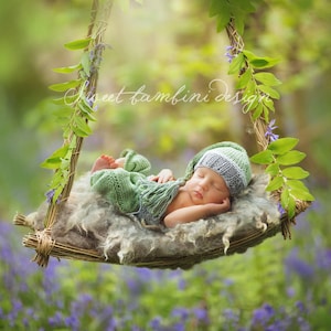 May include: A sleeping baby wearing a green and grey knitted hat lies in a woven basket swing decorated with green leaves and purple flowers. The swing is suspended from a branch in a woodland setting.