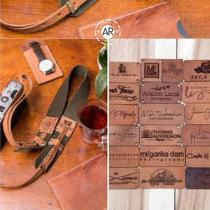 May include: A green leather camera strap with brown leather accents and metal rivets. The strap has several small leather patches with different text and logos, including "BitbyBit", "Avalon Lane Photography", "Thomas Auvrigon Photography", and "mriganka dam Photography".