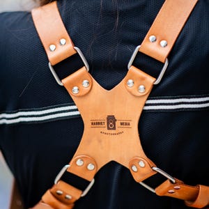 Pu&ograve; includere: Un'imbracatura per fotocamera in pelle marrone con hardware in metallo argentato. L'imbracatura ha un design a forma di X con il testo "HARRIET MEDIA & PHOTOGRAPHY" stampato sul davanti. L'imbracatura &egrave; indossata sopra una camicia nera.