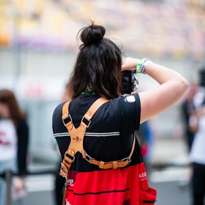 Pu&ograve; includere: Una persona indossa una maglietta nera e un grembiule rosso con la scritta "PHOTOGRAPHER" stampata sopra. Un'imbracatura per fotocamera in pelle marrone &egrave; indossata sopra la camicia.