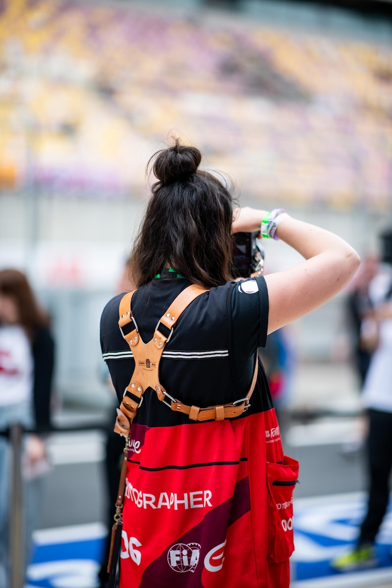 Pu&ograve; includere: Una persona indossa una maglietta nera e un grembiule rosso con la scritta "PHOTOGRAPHER" stampata sopra. Un'imbracatura per fotocamera in pelle marrone &egrave; indossata sopra la camicia.