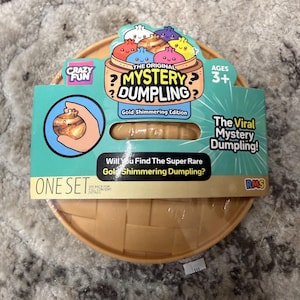 May include: A round, tan plastic container with a woven texture, featuring a cardboard label for The Original Mystery Dumpling Gold Shimmering Edition. The packaging displays colorful cartoon dumplings, text, and a cutout showing a tan toy dumpling.