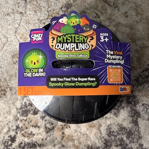 May include: A black circular container featuring a purple and orange cardboard label for the Mystery Dumpling Spooky Glow Edition. The label displays colorful cartoon dumplings, the text "Glow in the Dark!", a QR code, and the phrase "Will You Find The Super Rare Spooky Glow Dumpling?" for ages 3 and up.
