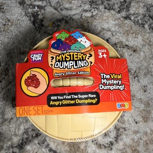 May include: A round, beige plastic dumpling steamer container featuring a red cardboard label that reads The Original Mystery Dumpling Angry Glitter Edition. The packaging includes illustrations of colorful dumplings and text asking Will You Find The Super Rare Angry Glitter Dumpling?
