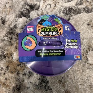 May include: A purple plastic container for The Original Mystery Dumpling Galaxy Edition toy. The packaging features cartoon dumplings, the text "Will You Find The Super Rare Galaxy Dumpling?", and "The Viral Mystery Dumpling!" with an age rating of 3+.