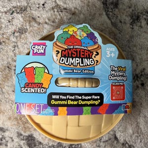 May include: A round, tan plastic container with a cardboard sleeve labeled The Original Mystery Dumpling Gummi Bear Edition. The packaging features colorful illustrations of dumplings and gummy bears, text reading Candy Scented, and a QR code.