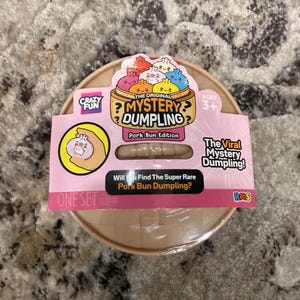 May include: A round, tan plastic container for The Original Mystery Dumpling Pork Bun Edition toy. The packaging features colorful cartoon dumplings, the text "Will You Find The Super Rare Pork Bun Dumpling?", and a "Crazy Fun" logo for ages 3+.