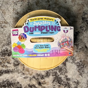 May include: A round, light yellow plastic container with a cardboard label featuring the text The Original Mystery Squishy Dumpling The Viral Fidget Toy Series 3. The label displays colorful dumpling illustrations and text about a super rare party confetti dumpling.
