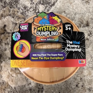 May include: A round, tan plastic container holding a mystery toy, featuring a black cardboard label with the text The Original Mystery Dumpling, Neon Edition, and The Viral Mystery Dumpling. The packaging includes colorful illustrations of dumplings.