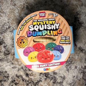 May include: A round cardboard packaging for a Mystery Squishy Dumpling toy. The label features the text "The Original Viral Mystery Squishy Dumpling" and "Rare Ruby Edition!" with colorful illustrations of smiling dumpling characters.