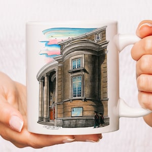 Puede incluir: Taza de cerámica blanca con una ilustración dibujada a mano de un edificio con columnas y una gran ventana. El edificio es marrón con una puerta roja y un letrero que dice "Convocation Hall, U of T". El cielo está pintado en tonos de azul, rosa y verde.