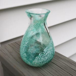 May include: A clear glass vase with a teal and white speckled design. The vase has a rounded body and a narrow neck. It is sitting on a weathered wooden surface.