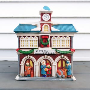 May include: A ceramic Christmas village station with a clock tower, blue roof, and the text "Victoria Station." The building features arched doorways with figures of people and a festive wreath. The station is painted in red, white, and green.