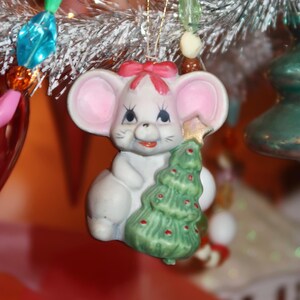 May include: A ceramic Christmas ornament shaped like a white mouse with pink ears and a red bow. The mouse is holding a green Christmas tree with red ornaments and a gold star. The ornament is hanging on a silver Christmas tree.