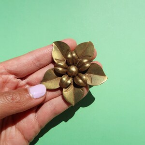 May include: A gold-toned metal flower brooch with five petals and a center detail. The brooch is held in a hand with a light pink nail polish.
