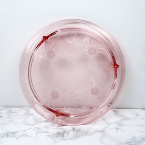 May include: A round, pink glass serving tray with a floral design. The tray has a raised edge and three small feet. The floral pattern includes stylized flowers and leaves. The tray is likely vintage and suitable for serving desserts.