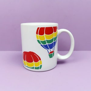 May include: White ceramic mug with a red, yellow, green, and blue hot air balloon design. The mug has a handle and is decorated with two hot air balloons.