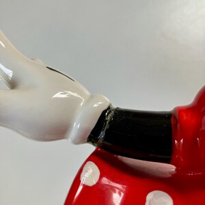 May include: Close-up of a ceramic figurine of a cartoon character wearing a red dress with white polka dots. The character's arm is outstretched with a white glove on the hand.