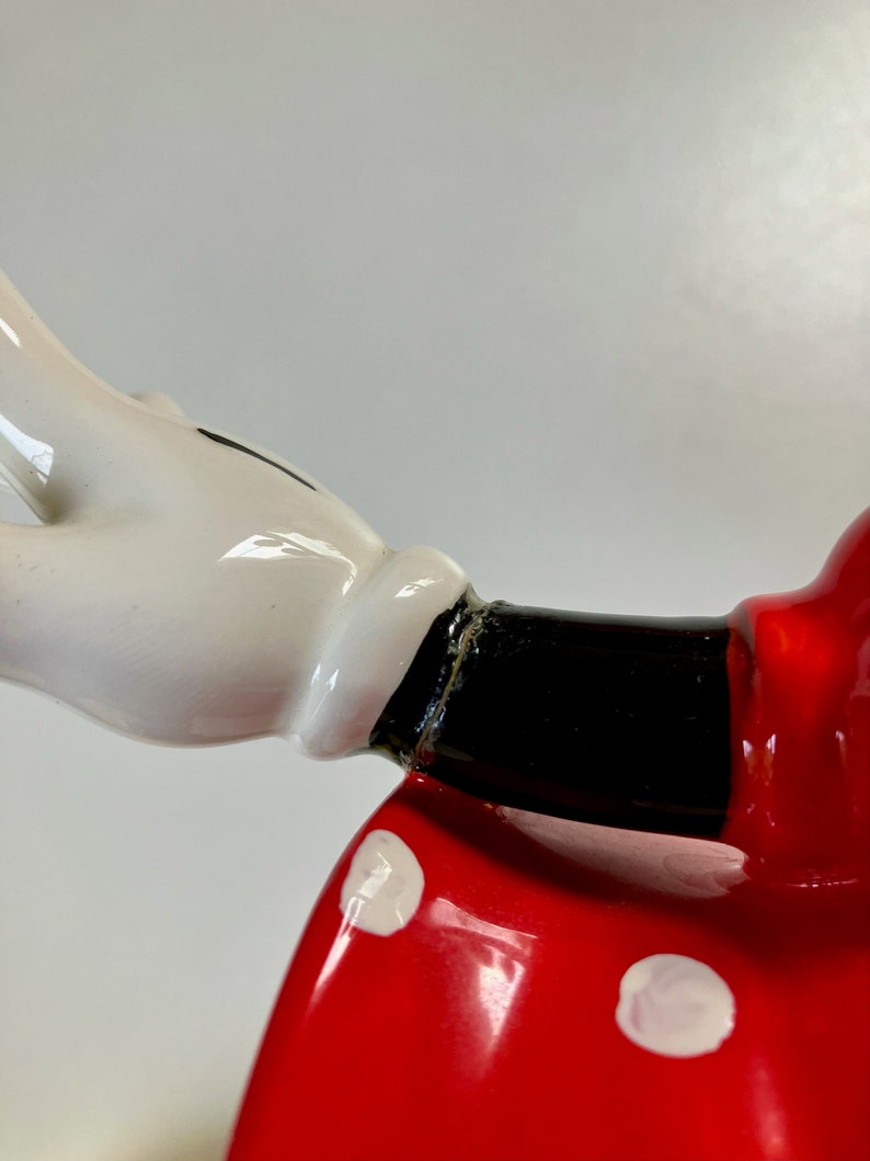 May include: Close-up of a ceramic figurine of a cartoon character wearing a red dress with white polka dots. The character's arm is outstretched with a white glove on the hand.