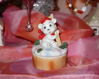 May include: A ceramic Christmas-themed trinket box featuring a white polar bear wearing a red hat and scarf, holding a wooden ski pole. The bear sits atop a snow-covered lid with holly accents, on a light brown base.