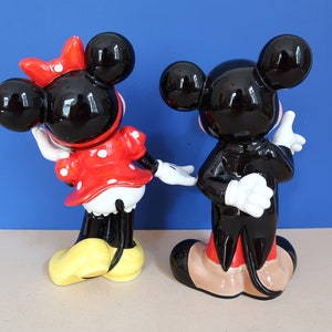 May include: A black and white ceramic figurine of Mickey Mouse wearing a tuxedo, standing next to a black and white ceramic figurine of Minnie Mouse wearing a red and white polka dot dress.