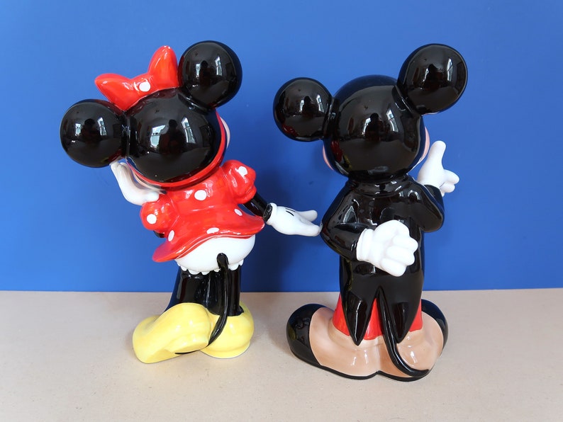 May include: A black and white ceramic figurine of Mickey Mouse wearing a tuxedo, standing next to a black and white ceramic figurine of Minnie Mouse wearing a red and white polka dot dress.