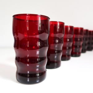May include: A set of eight red glass tumblers. The glasses have a unique, wavy design and are arranged in a diagonal line against a white background. The color of the glasses is a deep, rich red.