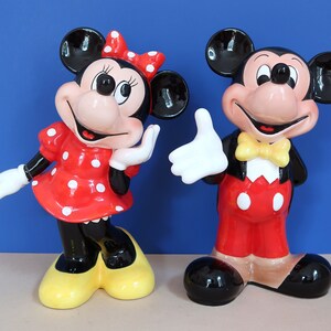 May include: A pair of ceramic Mickey Mouse and Minnie Mouse figurines. Mickey is wearing a red and white outfit with a yellow bow tie. Minnie is wearing a red and white polka dot dress with a red bow. Both figurines are standing on a light brown surface.