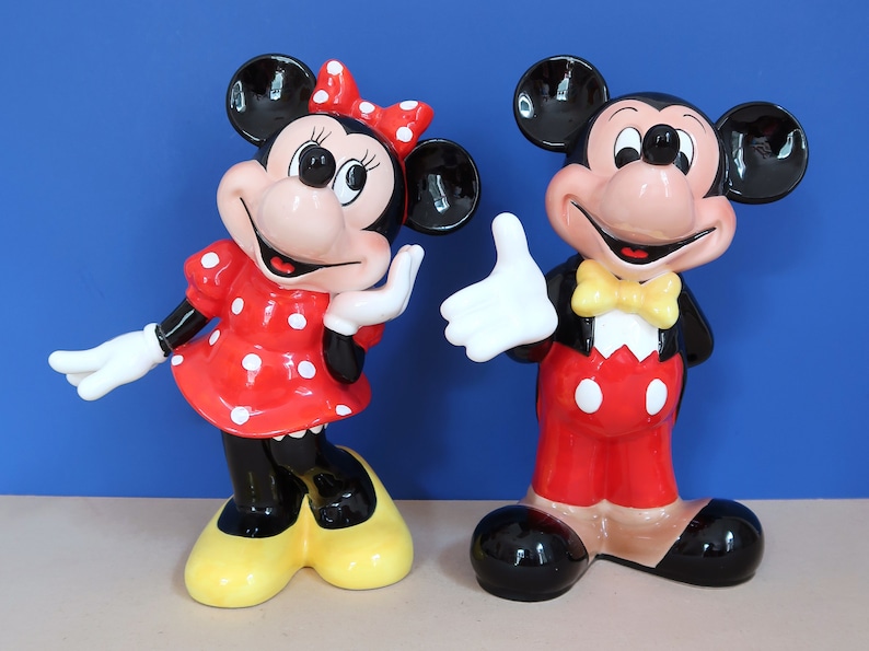 May include: A pair of ceramic Mickey Mouse and Minnie Mouse figurines. Mickey is wearing a red and white outfit with a yellow bow tie. Minnie is wearing a red and white polka dot dress with a red bow. Both figurines are standing on a light brown surface.