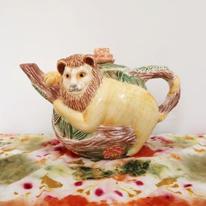 May include: A ceramic teapot featuring a lion design. The teapot has a light yellow body with a lion's head and mane, brown branches, and green leaves. The handle and spout are brown. The teapot is sitting on a colorful patterned surface.
