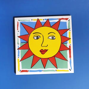 May include: Square ceramic tile featuring a stylized sun design. The sun has a yellow face with red rays, a red mouth, and black eyes. The tile is bordered by a colorful geometric pattern.