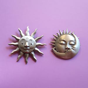 May include: Two silver-toned celestial-themed wall hangings. One is a smiling sun with radiating rays, the other a crescent moon with a face. Both have a vintage, antique appearance. Set against a purple background.