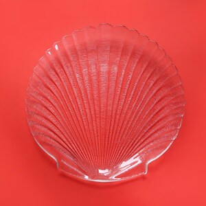 May include: Clear glass seashell-shaped plate with a textured surface. The plate has a scalloped edge and a fan-like pattern radiating from the center. The plate is set against a solid red background.