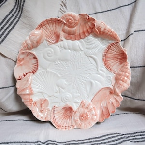 May include: A white and coral-colored ceramic plate with a seashell design. The plate features raised relief seashell shapes around the rim and in the center. The plate is round and has a scalloped edge.