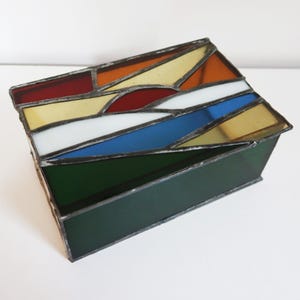 May include: A stained glass box with a geometric design. The lid features a colorful pattern of red, orange, yellow, white, and blue glass pieces. The box's sides are a dark green color.