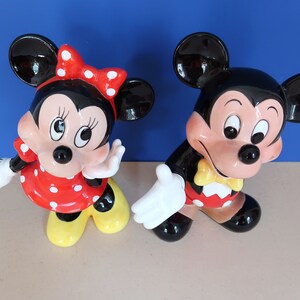 May include: A pair of ceramic Mickey Mouse and Minnie Mouse figurines. Mickey is wearing a red and yellow outfit with a yellow bow tie. Minnie is wearing a red and white polka dot dress with a red bow.
