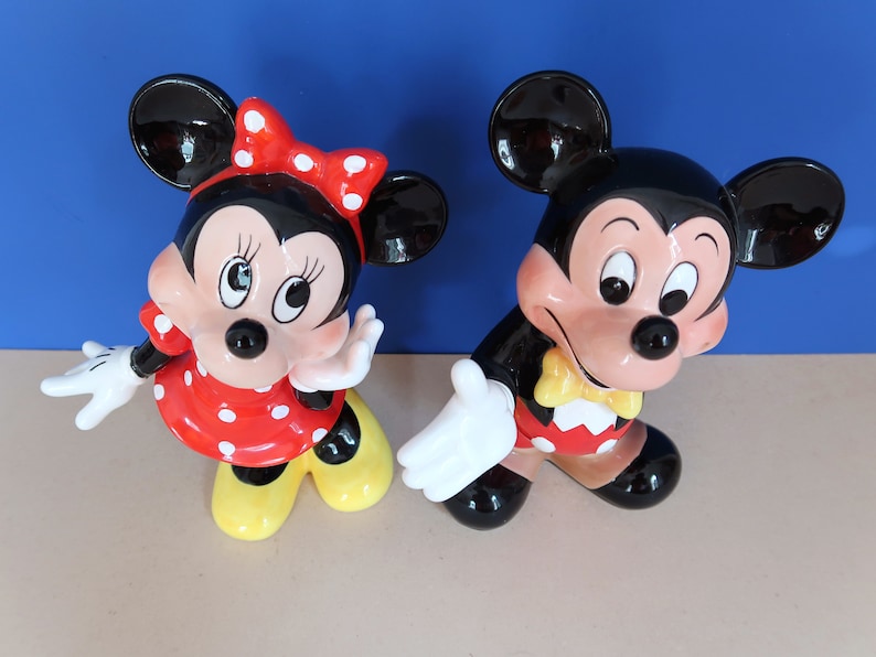 May include: A pair of ceramic Mickey Mouse and Minnie Mouse figurines. Mickey is wearing a red and yellow outfit with a yellow bow tie. Minnie is wearing a red and white polka dot dress with a red bow.