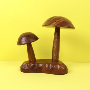 May include: A wooden mushroom-shaped decorative item. The item features two mushrooms of different sizes, with rounded caps and stems, connected by a carved base. The wood has a warm, brown tone, and the background is yellow.