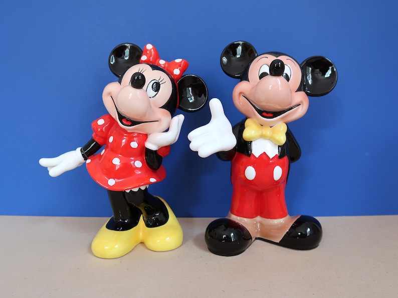 May include: A pair of ceramic figurines of Mickey Mouse and Minnie Mouse. Mickey is wearing a red and black tuxedo with a yellow bow tie. Minnie is wearing a red dress with white polka dots and a yellow bow.