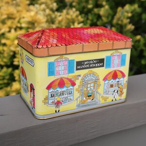 May include: A rectangular metal tin featuring a yellow exterior with illustrations of a storefront labeled Ye Olde Sweet Shoppe. The lid is painted with a red and orange shingled roof design. The sides depict windows, blue shutters, and figures.