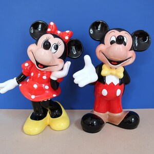 May include: A pair of ceramic figurines of Mickey Mouse and Minnie Mouse. Mickey is wearing a red and white outfit with a yellow bow tie. Minnie is wearing a red and white polka dot dress with a red bow. Both figurines have black shoes.