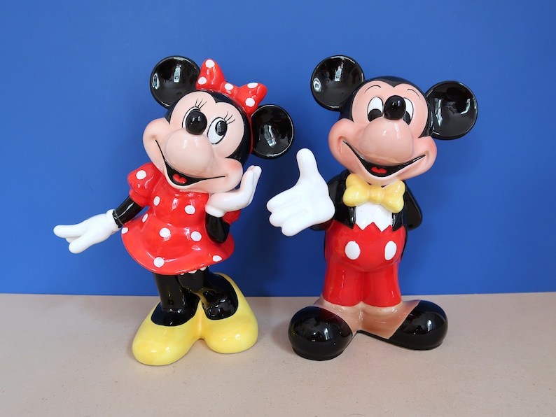 May include: A pair of ceramic figurines of Mickey Mouse and Minnie Mouse. Mickey is wearing a red and white outfit with a yellow bow tie. Minnie is wearing a red and white polka dot dress with a red bow. Both figurines have black shoes.